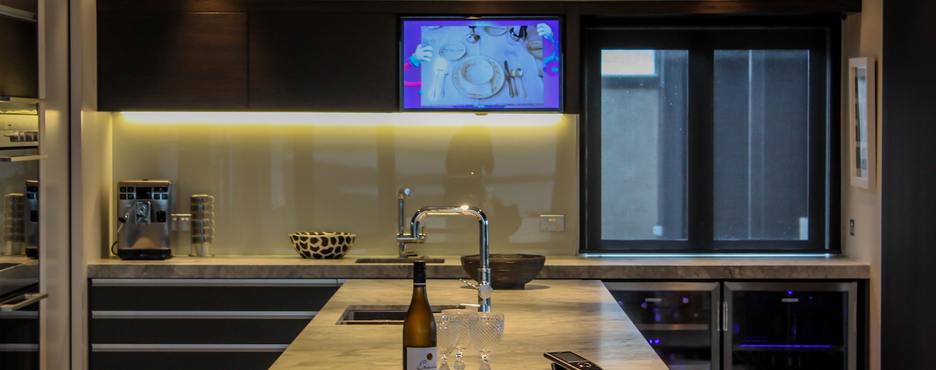 Modern kitchen with TV screen in cabinetry over bench controlled through home automation remote on bench