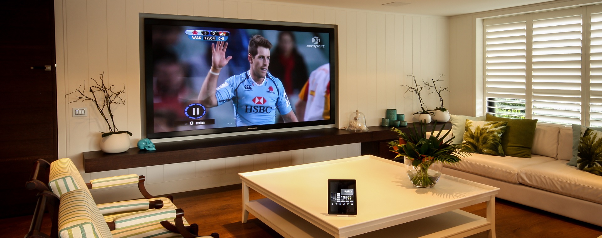 Modern lounge with large display screen showing Rugby League on screen, and touchscreen control tablet on coffee table