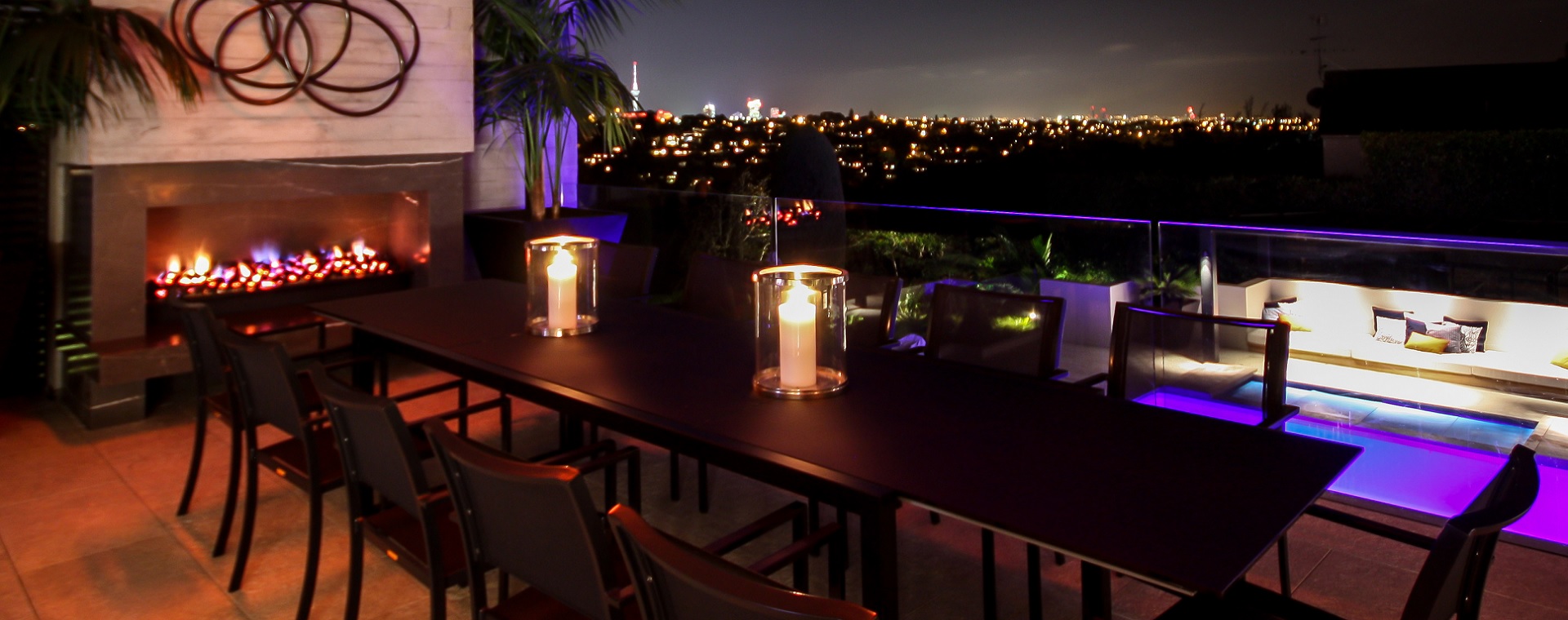 Stunning outdoor dining setting with fireplace, pool and evening cityscape featuring home automation control