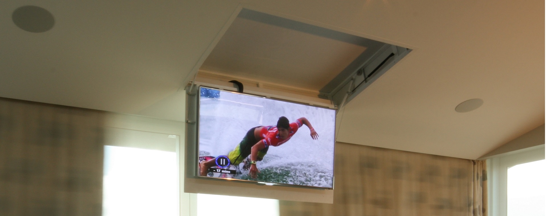 TV screen on a screen dropper showing a surfer, in a bedroom with in-ceiling speakers