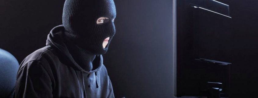 Man in black balaclava, hoodie and gloves hacking a computer
